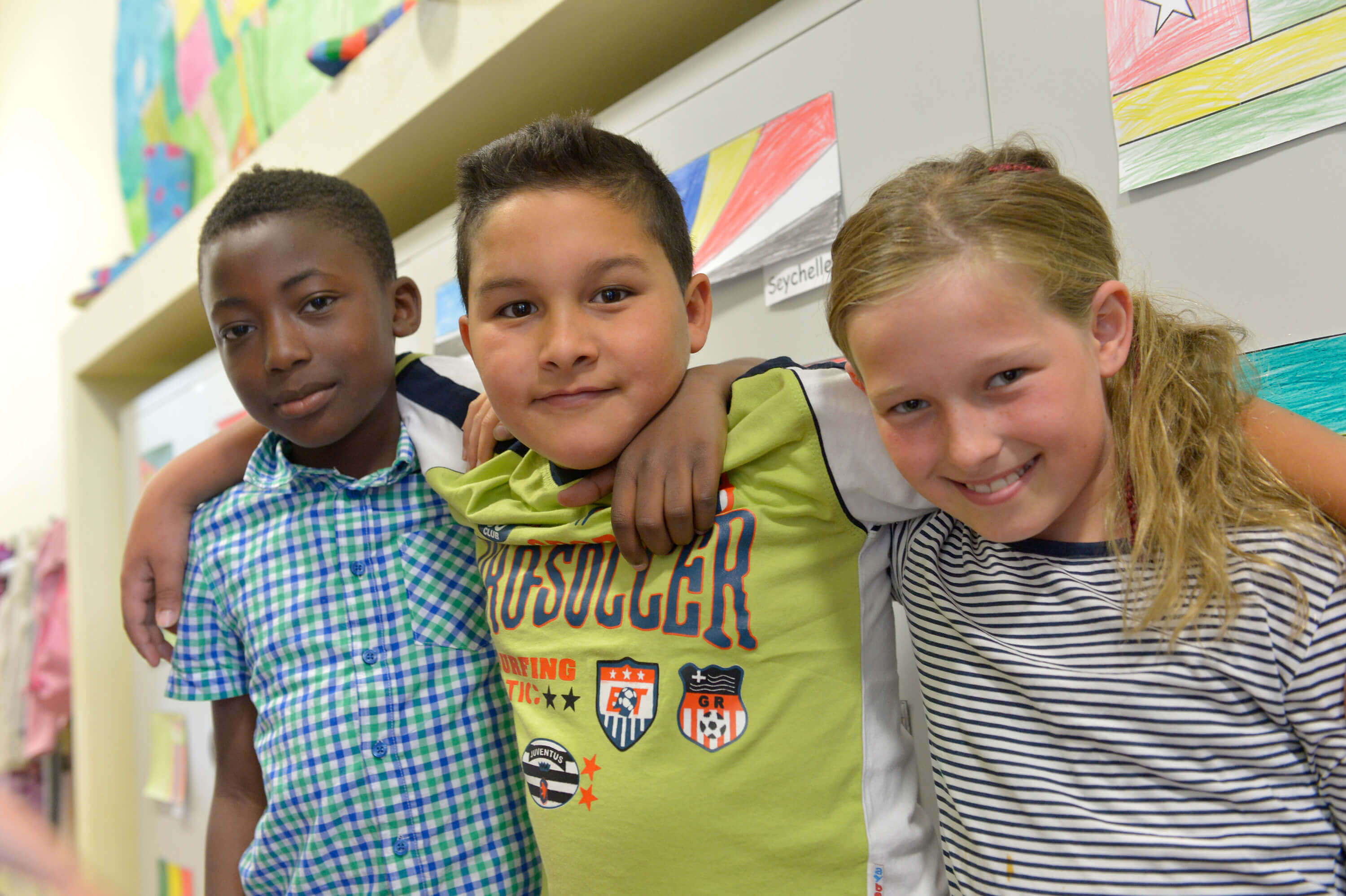 drei Kinder im Grundschulalter, die ihre Arme umeinander legen, ein Junge mit dunkler Hautfarbe, ein Junge mit dunklen Haaren, ein blondes Mächen